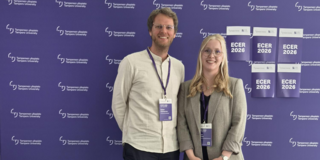 Ruben Kleinkorres und Laura Becher stehen vor einer blauen Wand mit dem Tagungslogo