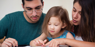 Eltern (ein Mann und eine Frau) lesen ihrer Tochter aus einem Buch, welches vor ihnen liegt, vor.