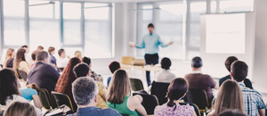 Foto, welches einen hellen Konferenzraum zeigt, wo ein Sprecher mit blauem Hemd eine Präsentation vor Publikum hält.