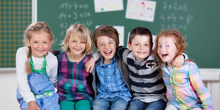 Fünf Grundschulkinder vor einer Schultafel umarmen sich und lächeln in die Kamera