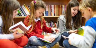 Vier Grundschülerinnen und Grundschüler sitzen vor einem Bücherregal am Boden und lesen gemeinsam in Büchern