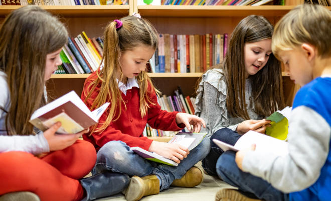 Vier Grundschülerinnen und Grundschüler sitzen vor einem Bücherregal am Boden und lesen gemeinsam in Büchern