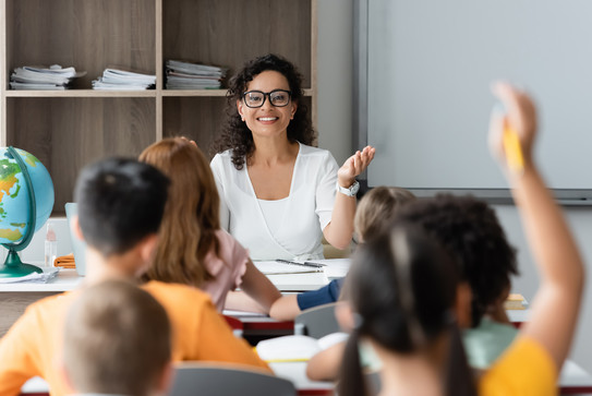 Lehrkraft lächelt zu Grundschulkindern, die ihre Hände zur Wortmeldung in die Luft strecken