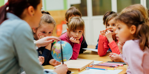 Kinder sitzen um einen Glous herum, der von der Lehrerin in der Hand gehalten wird