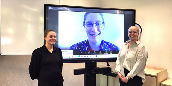 Carina Jüschke und Laura Becher vor einem großen Bildschirm, auf dem Dr. Katherine Reynolds zugeschaltet zu sehen ist.