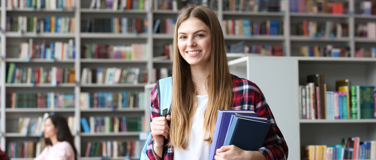 Eine lächelnde Studentin mit Büchern in einem Arm vor mehreren Bücherregalen