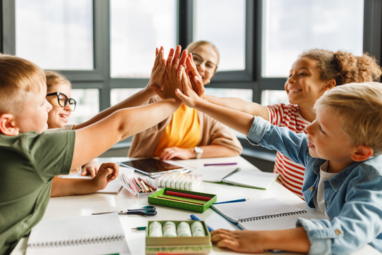 Grundschulkinder und eine Lehrkraft sitzen gemeinsam am Tisch im Klassenraum und legen die Hände zusammen