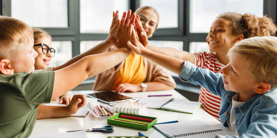 Grundschulkinder und eine Lehrkraft sitzen gemeinsam am Tisch im Klassenraum und legen die Hände zusammen