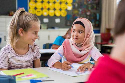 Zwei Schülerinnen, wovon eine die Hijab trägt, unterhalten sich am Tisch im Klassenraum