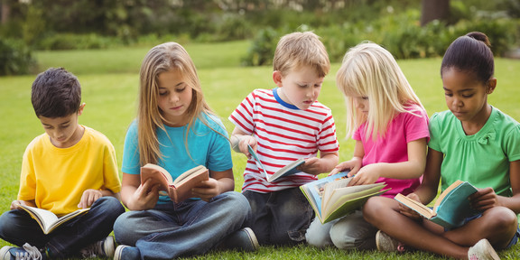 Fünf Kinder sitzen auf einer Wiese in einem Park und lesen gemeinsam Bücher.