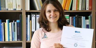 Theresa Schlitter steht mit ihrer JURE Urkunde vor einem Bücherregal