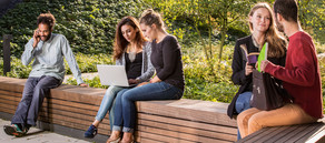 Mehrere Studierende sitzen auf einer langen Holzbank und unterhalten sich miteinander oder arbeiten am Laptop.