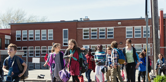 Schulschluss Mehrere Schülerinnen und Schüler laufen über den Zebrastreifen einer Straße vor ihrem Schulgebäude