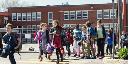 Mehrere Schülerinnen und Schüler laufen über den Zebrastreifen einer Straße vor ihrem Schulgebäude