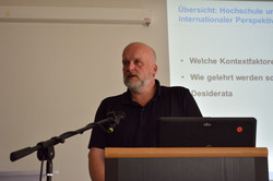 Foto von Prof. Dr. Holger Horz am Pult mit Mikrophon und Powerpointfolie im Hintergrund