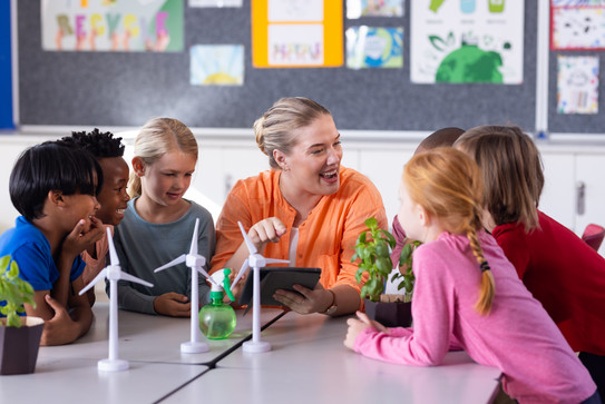 Lehrkraft sitzt mit mehreren Grundschulkindern am Tisch und bespricht Windkraft mit Modellen von Windmühlen