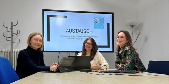 Drei Forscherinnen sitzen an einem Tisch, im Hintergrund ein Bildschirm mit einer Präsentation mit dem Titel "Austausch"