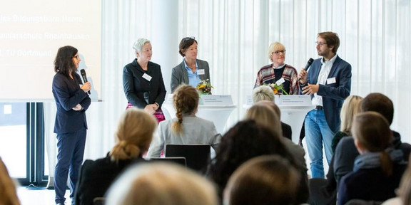 Fünf Personen stehen vor einer größeren Anzahl von Menschen und halten eine Podiumsdiskussion 