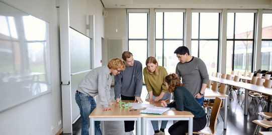 Personen arbeiten in einem Seminarraum in einer Gruppe zusammen, auf dem Tisch steht das TU-Logo.
